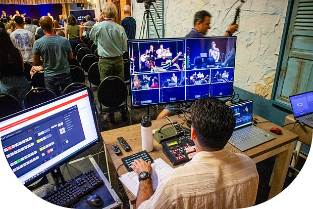 A video production operator manages a multi-camera live event setup, with several monitors showing camera feeds while an audience watches a presentation in the background.