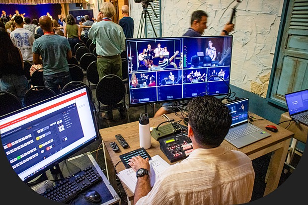 A video production operator manages a multi-camera live event setup, with several monitors showing camera feeds while an audience watches a presentation in the background.